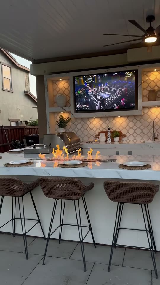 Marble bar island with burning fire trough, grill and mounted TV above arabesque tile at dusk