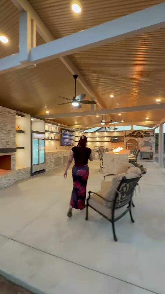 Woman walking through a large gable-covered outdoor kitchen with stone fireplace, TV and fire-trough bar