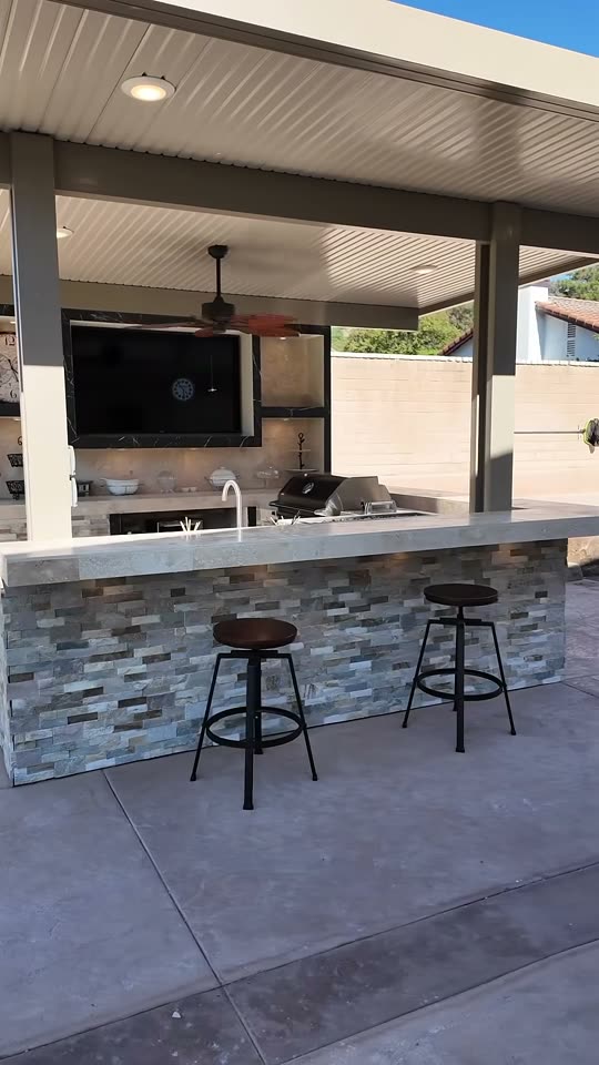 Stacked-stone kitchen bar with grill and sink under an alumawood patio cover, marble TV wall behind