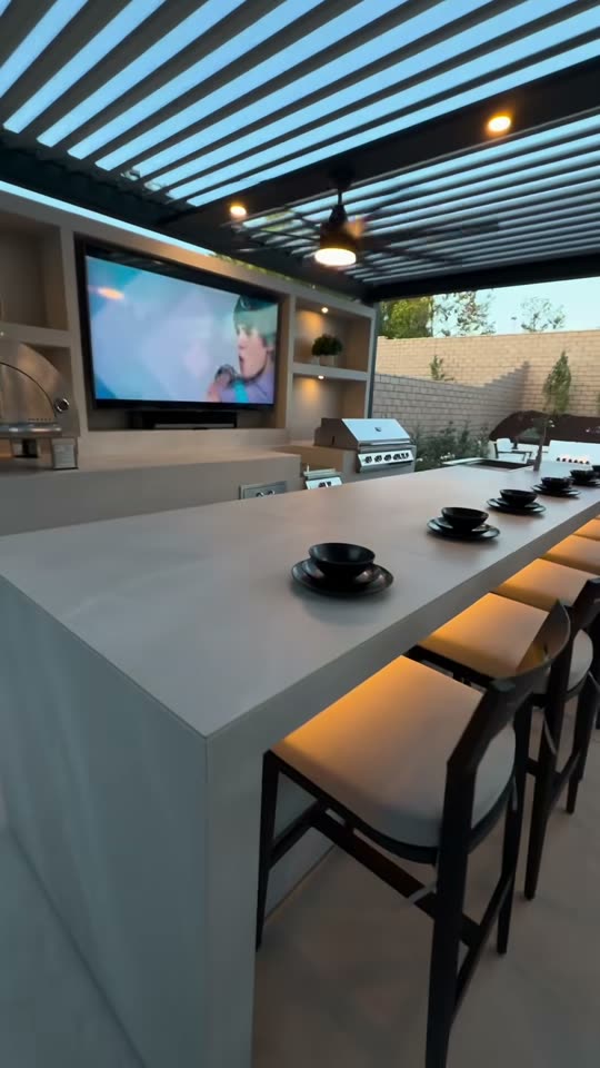 Concrete bar counter set for dinner under a louvred pergola, with stainless grill and mounted TV