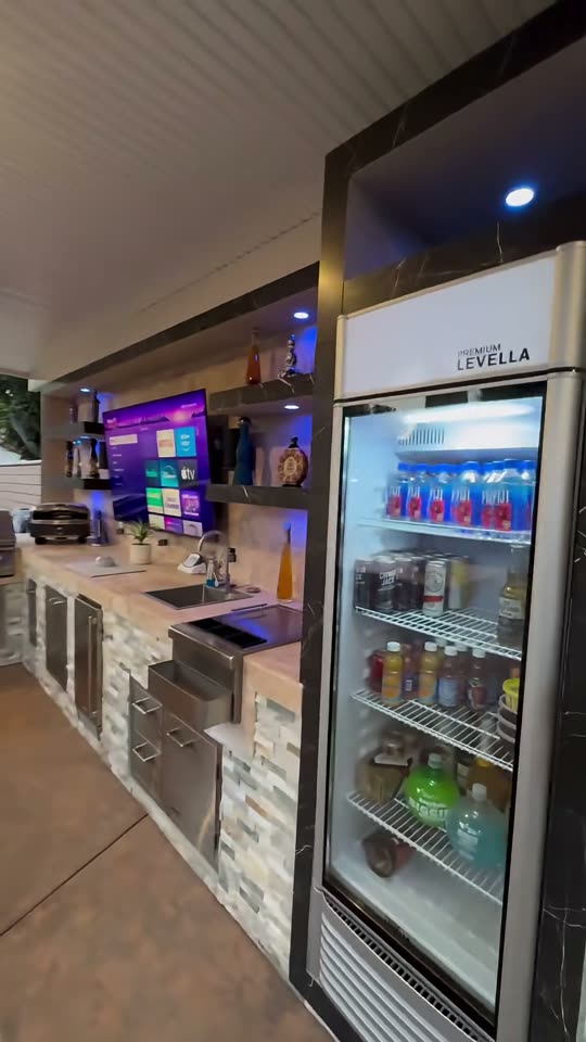 Stone bar with marble-framed shelving, mounted TV and a tall glass-door beverage cooler at night