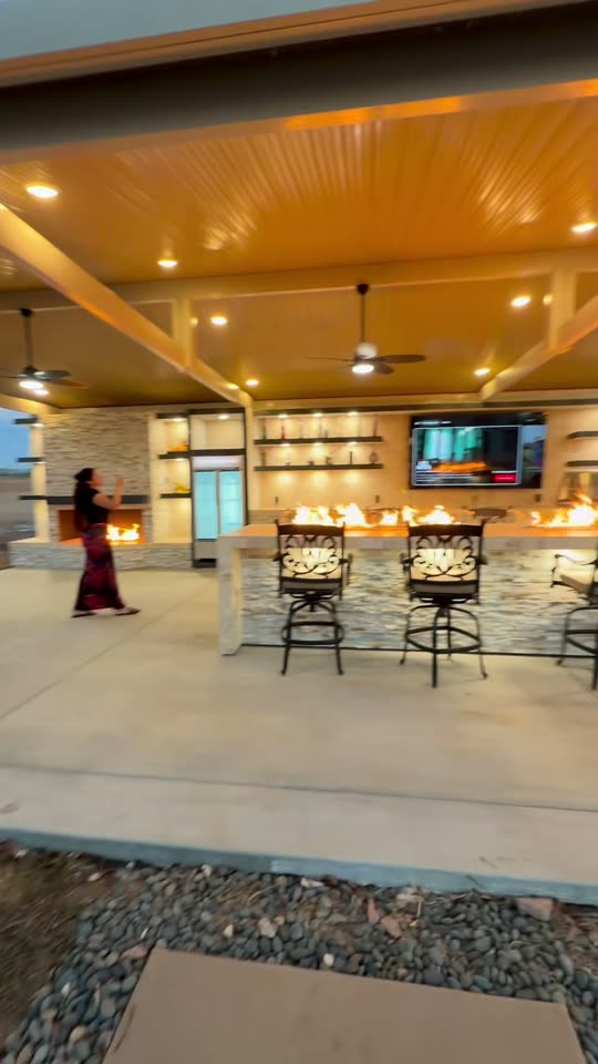 Blurred dusk pan across a burning fire-trough bar and mounted TV under a wide gable patio cover
