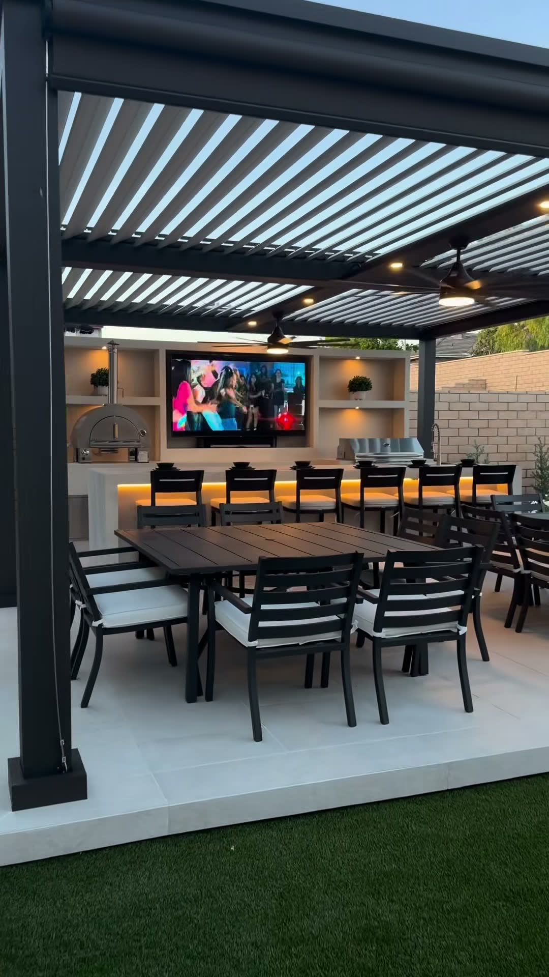 Black louvred aluminium pergola over a dining patio with a backlit bar, TV wall and pizza oven at dusk
