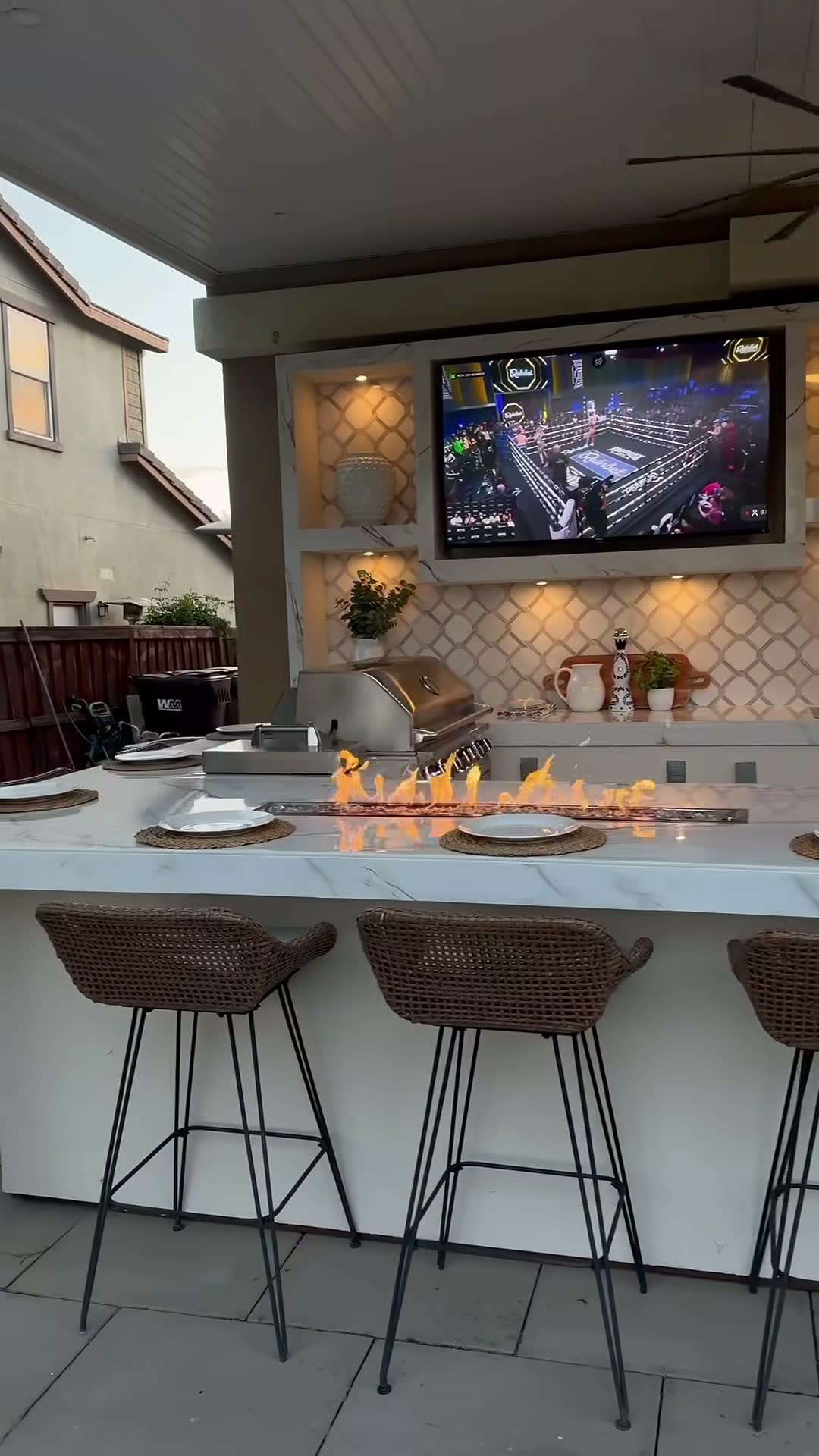 Marble-topped outdoor kitchen bar with a burning linear fire trough, stainless grill and mounted TV above arabesque tile