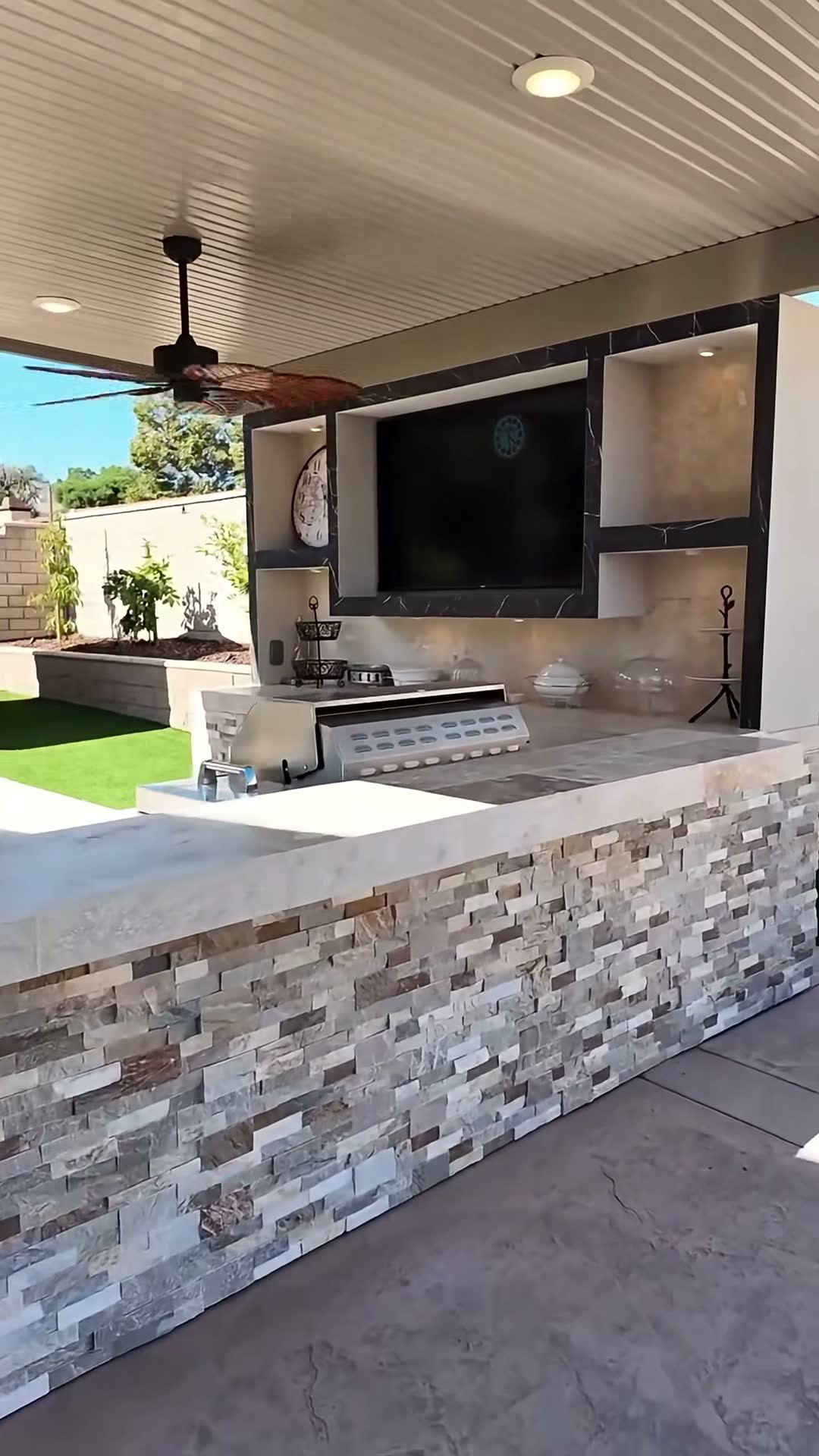Stacked-stone outdoor kitchen with stainless grill and sink under a solid patio cover, black marble TV wall behind