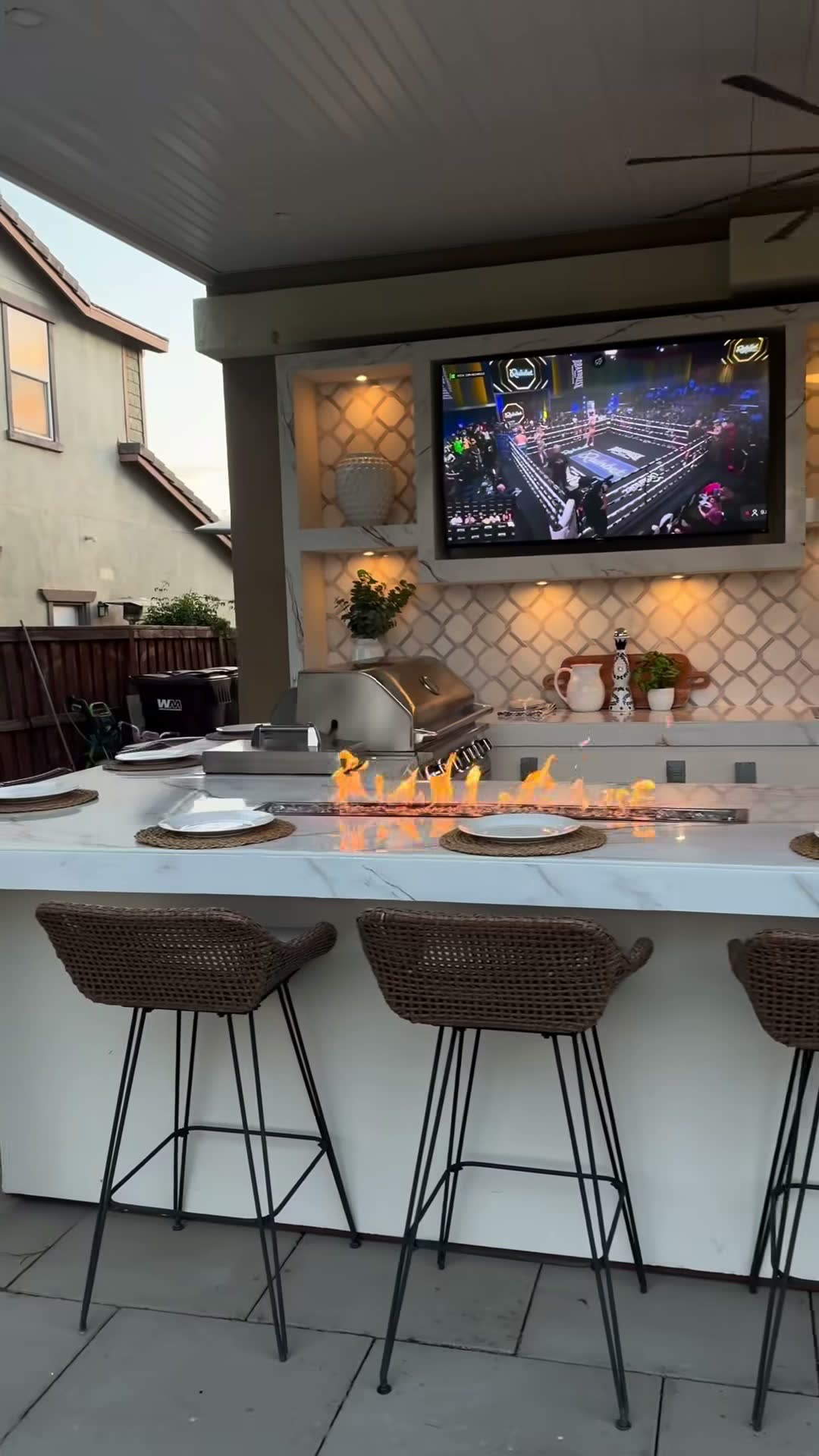 Fire trough burning along a white marble bar top beside a stainless grill, TV mounted on tiled wall