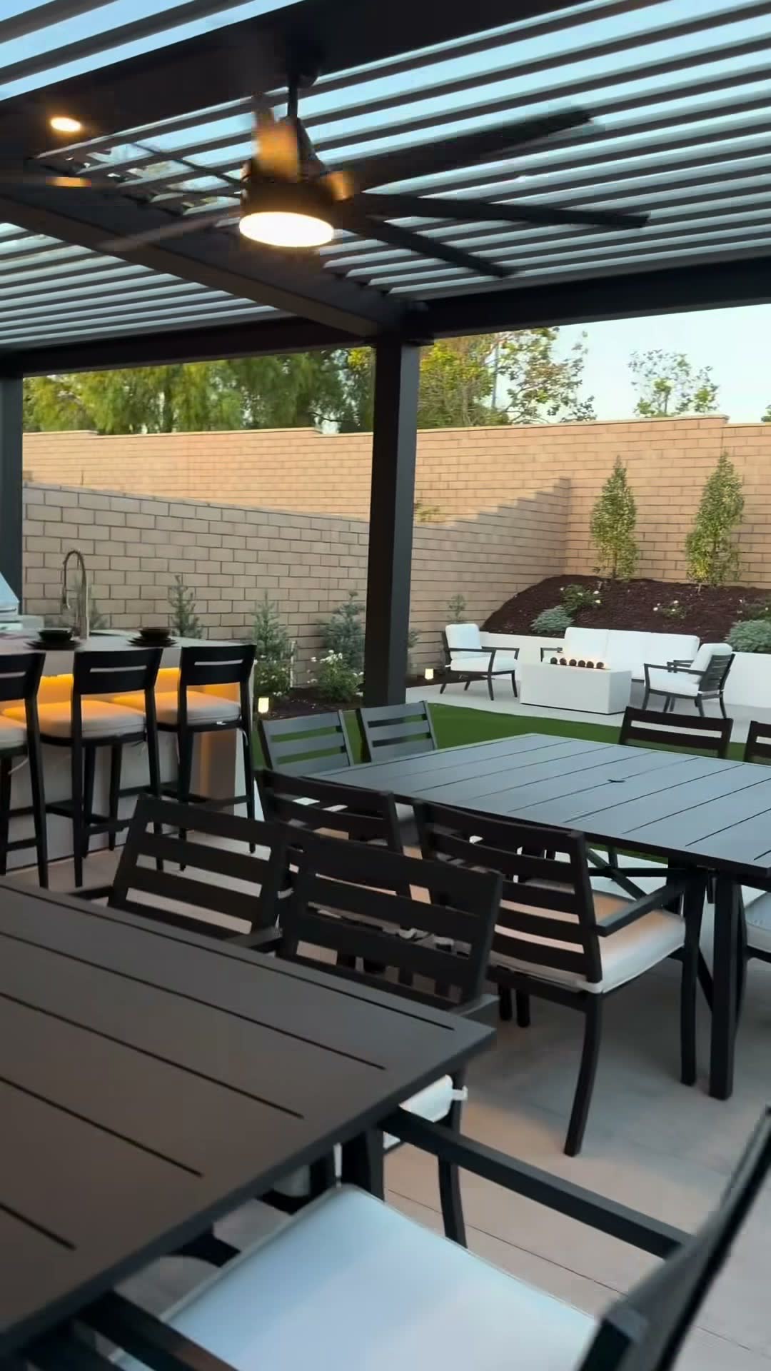 View from under a black louvred pergola toward a lit linear fire table and white lounge seating
