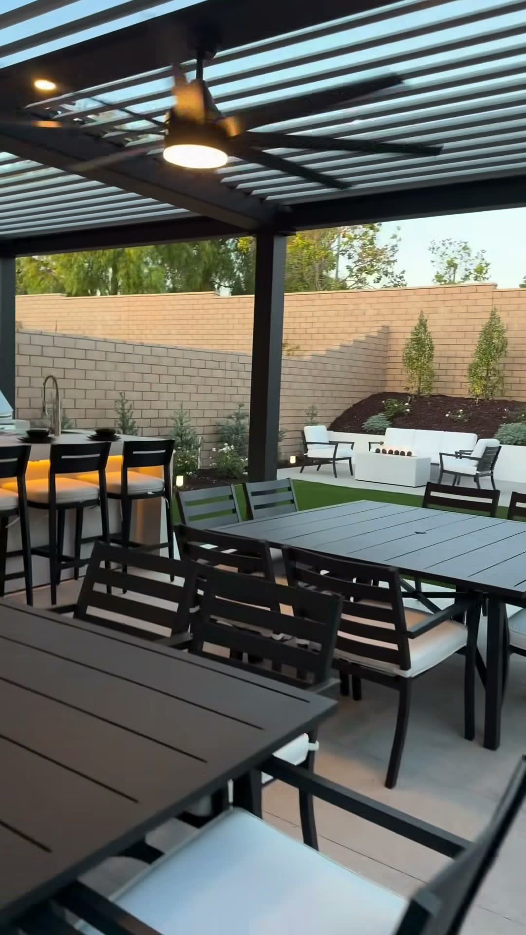 Louvred pergola over patio dining tables, with a gas fire table and lounge set on the lawn beyond