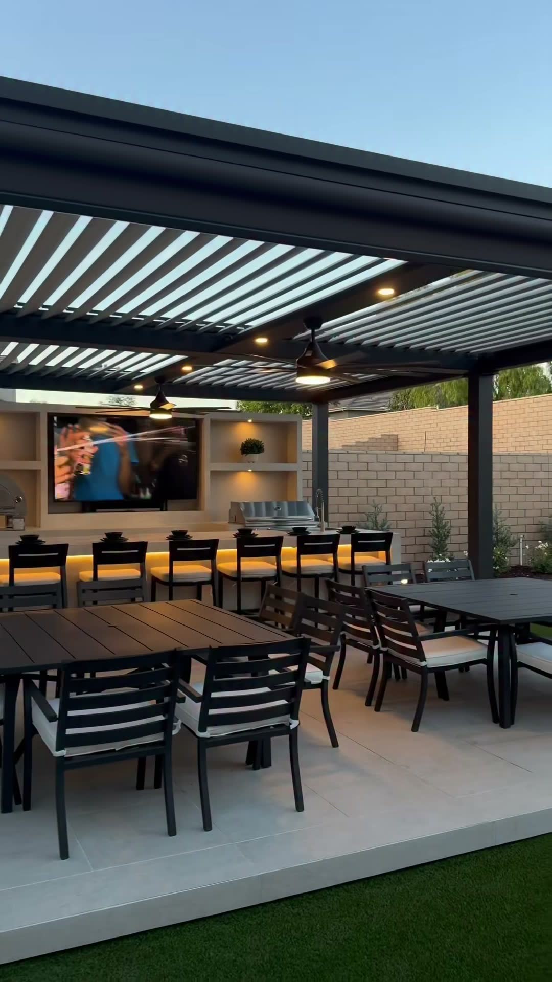 Wide dusk view of a louvred pergola sheltering a backlit bar run, stainless grill and mounted TV wall
