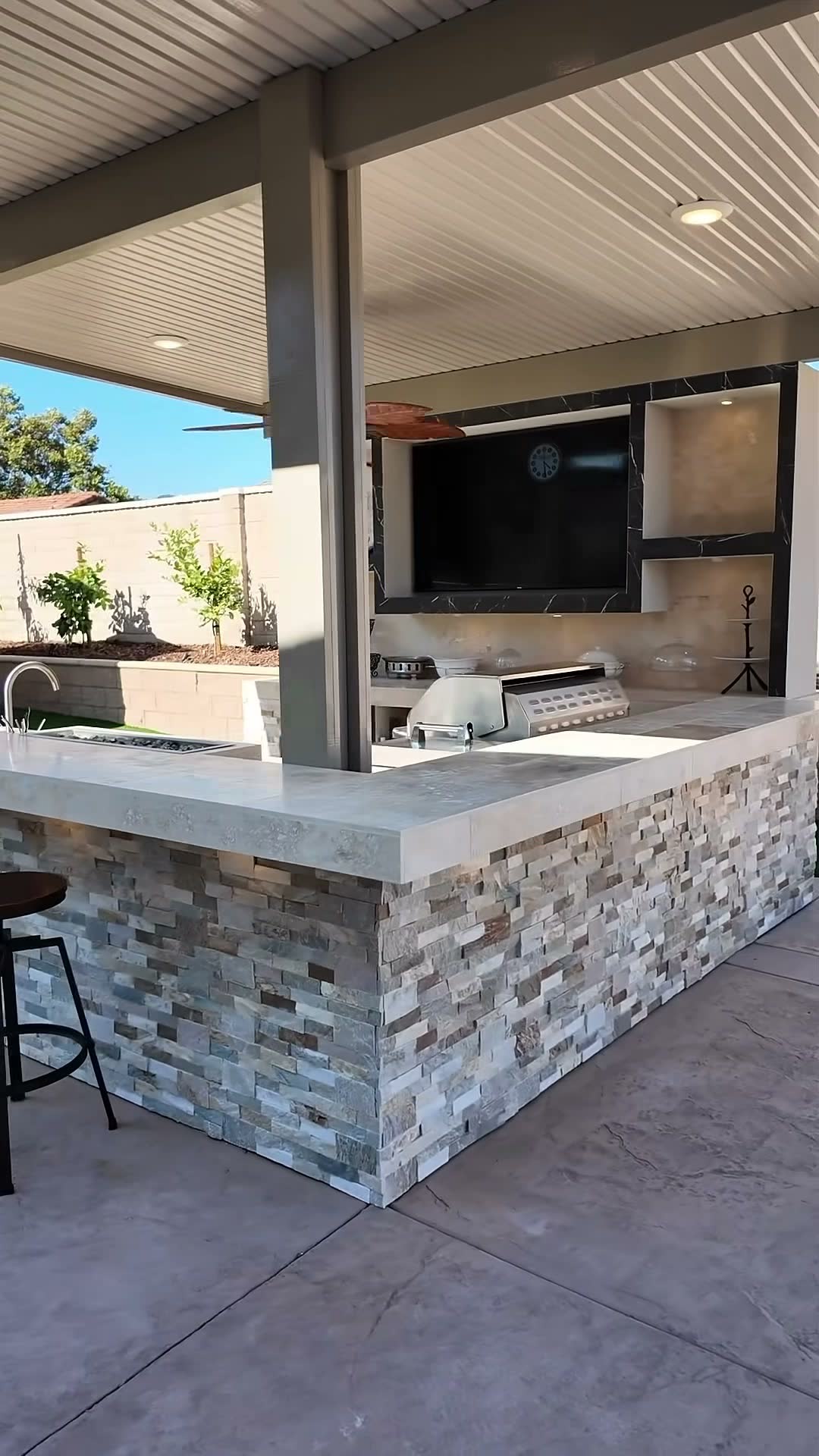 Corner view of an L-shaped stacked-stone bar with sink and grill under a solid patio cover
