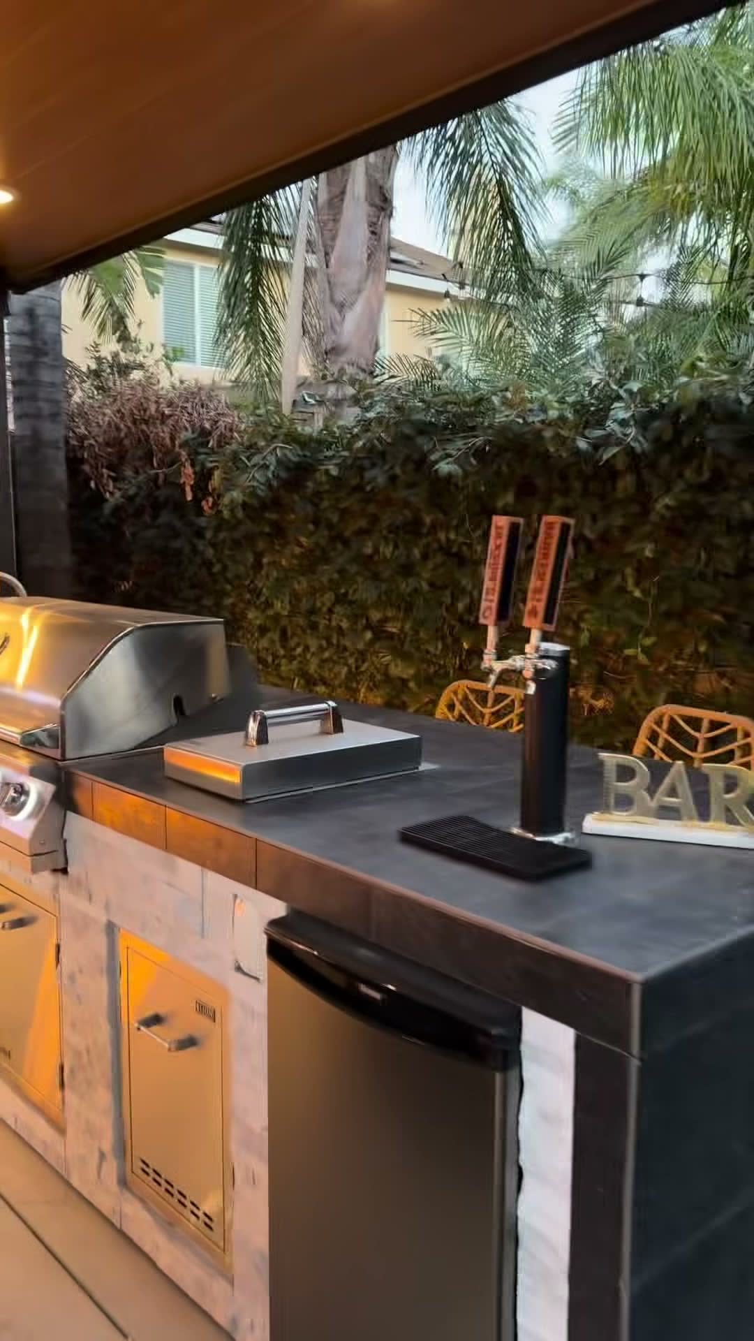 BBQ island with dark counter, stainless grill and a twin-tap kegerator under a wood-clad patio cover