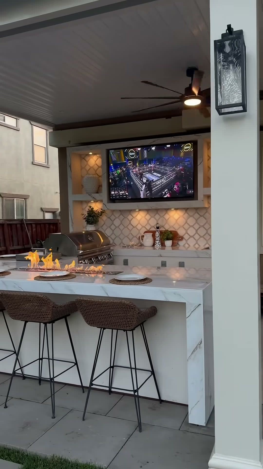 White marble bar island with a burning fire trough, stainless grill and mounted TV under a patio cover