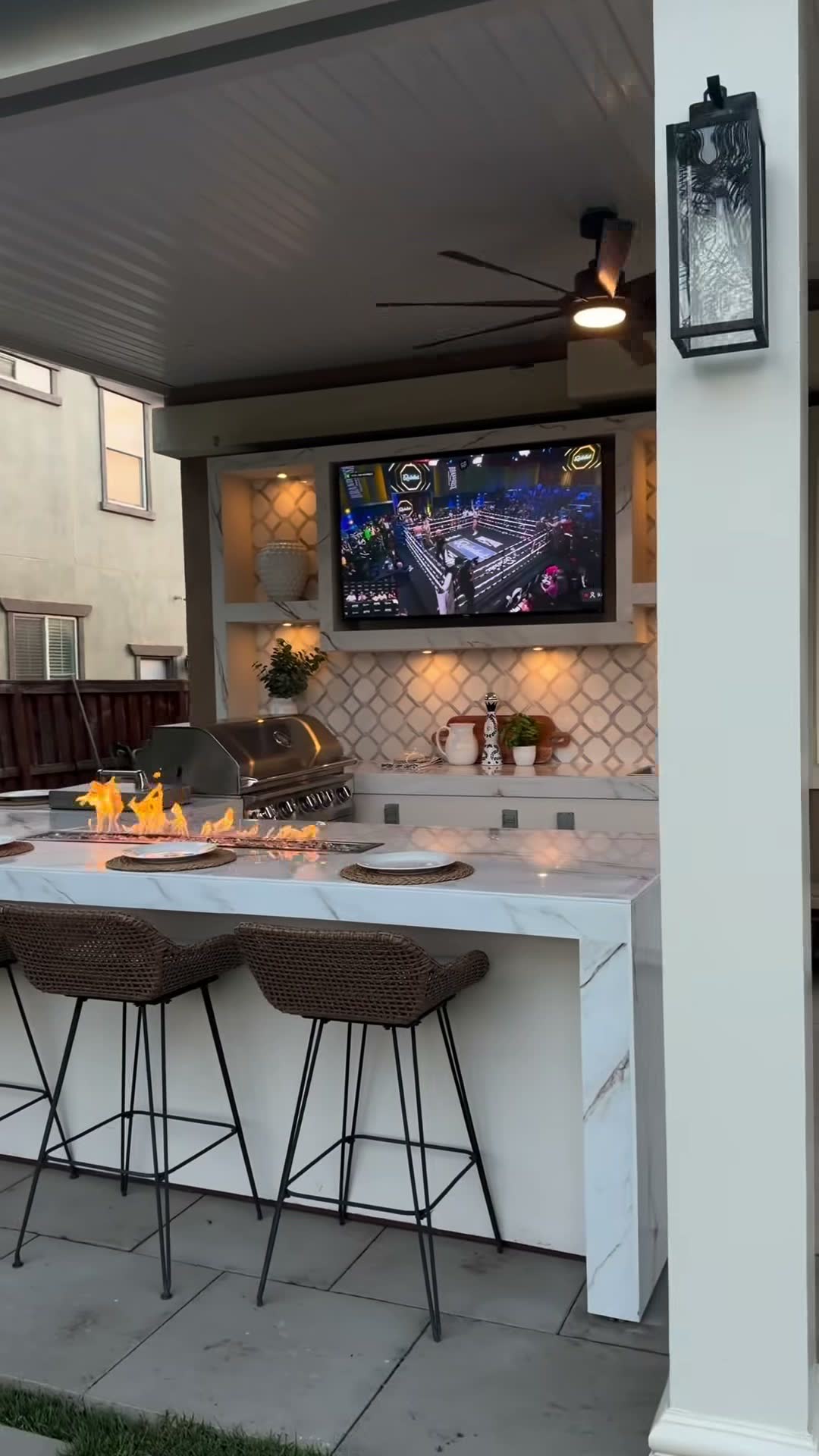 Marble bar with lit fire trough and wicker stools, TV mounted above a tiled backsplash and grill