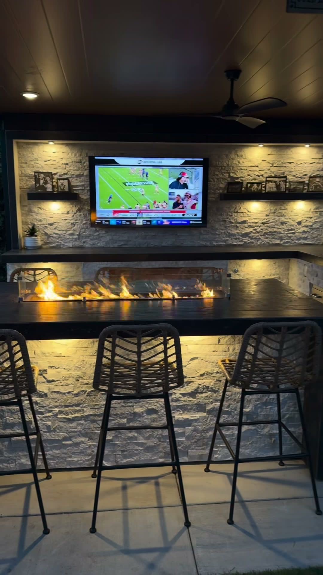 Stacked-stone TV wall with lit floating shelves above a bar-top fire trough and rope stools at night