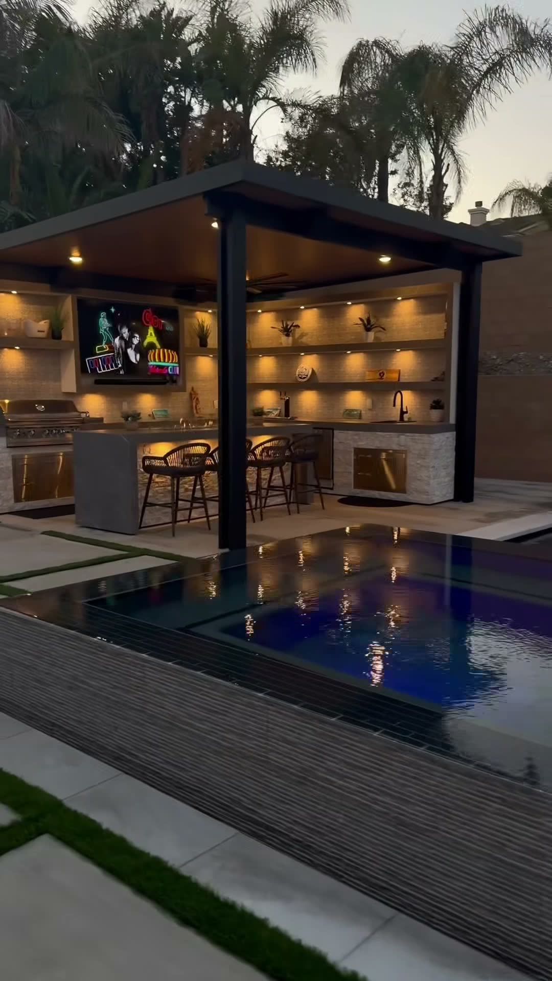Poolside bar under a black flat-roof cabana with mounted TV, backlit shelving and stainless grill at dusk