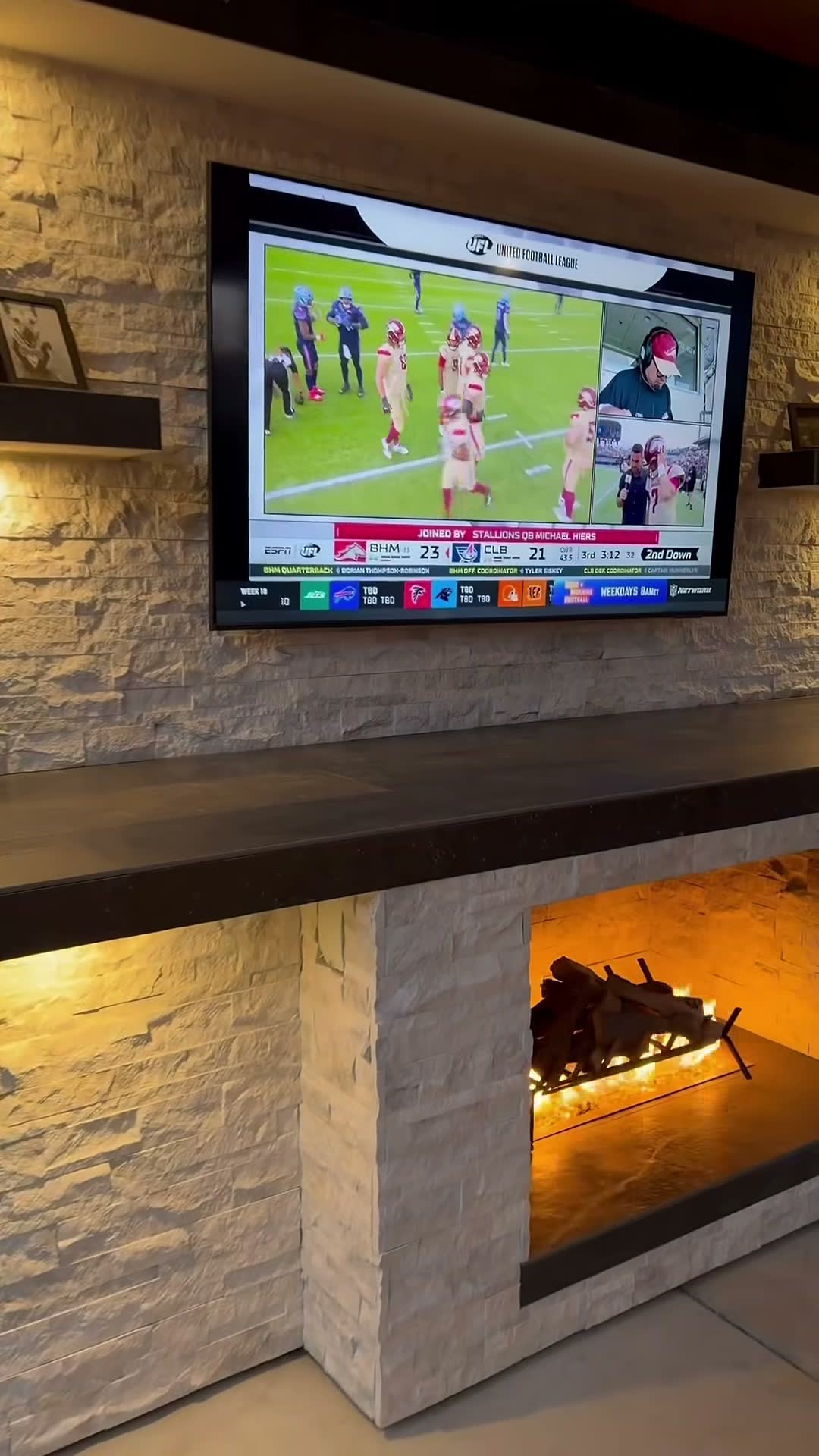 Mounted TV on backlit stacked stone above a see-through gas log fireplace at night