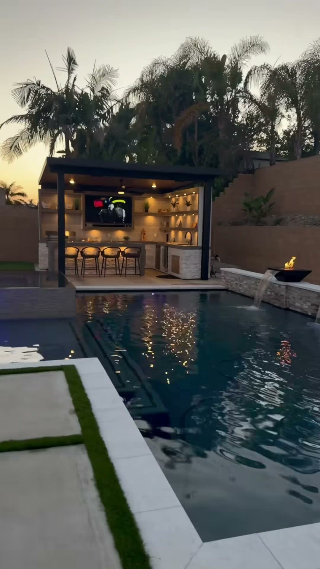 Sunset view across a pool to a lit cabana bar with mounted TV, plus a stone fire bowl and scupper