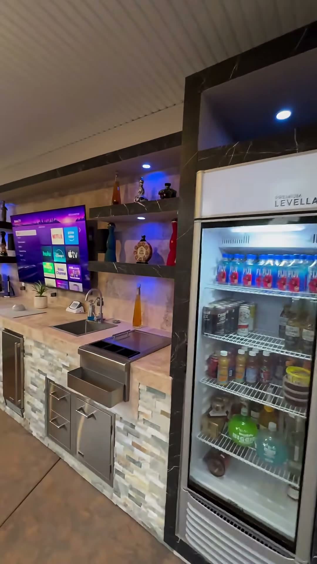 Night view of a stone bar run with mounted TV, LED-lit marble shelving and a glass-door beverage cooler