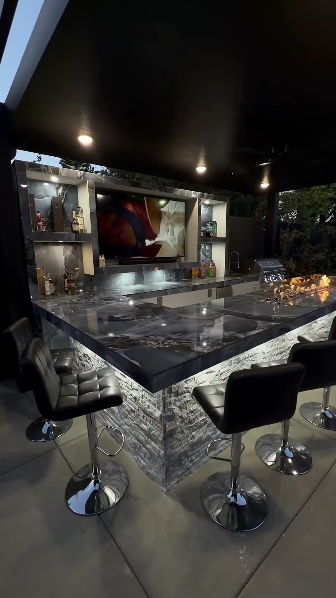 L-shaped bar in dark marble with backlit stone base, bar-top fire trough, grill and mounted TV