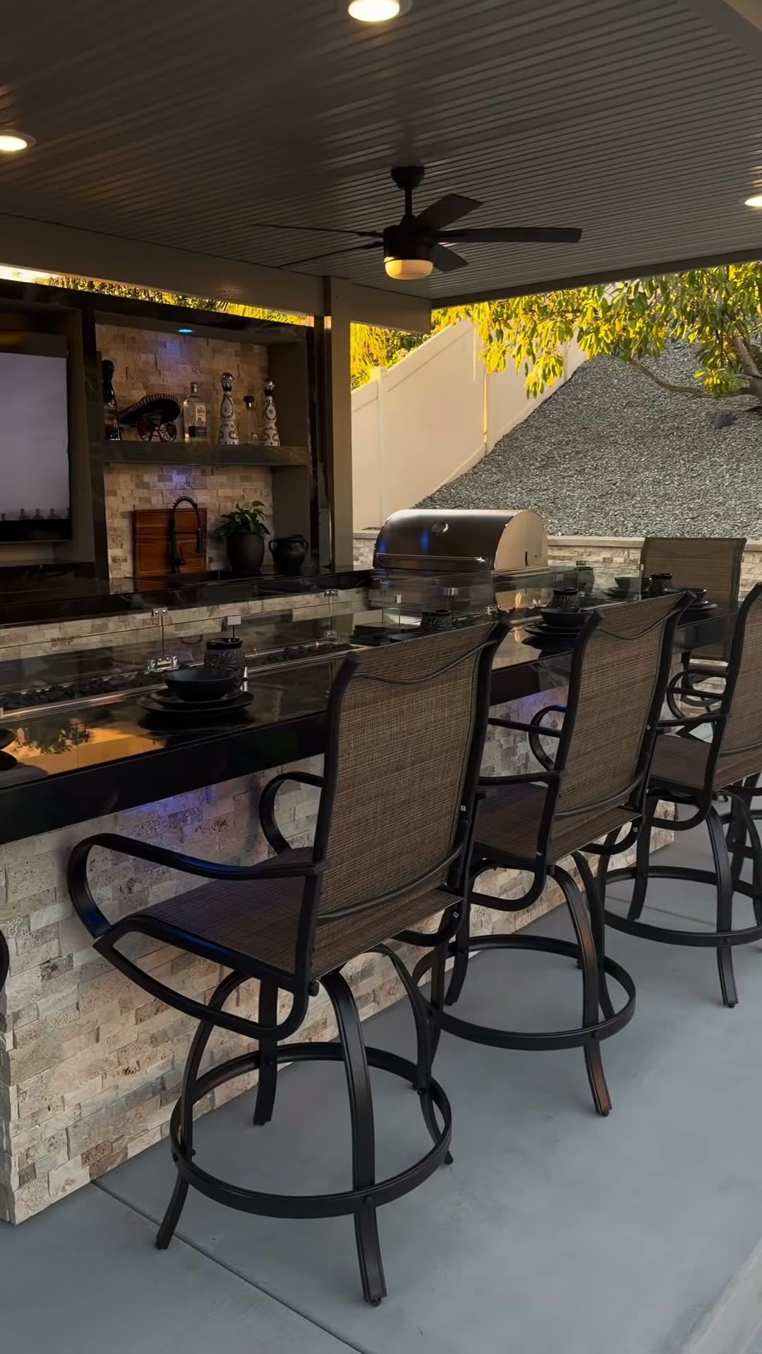 Long travertine bar with glass-shielded fire trough, grill and lit TV wall under an alumawood cover