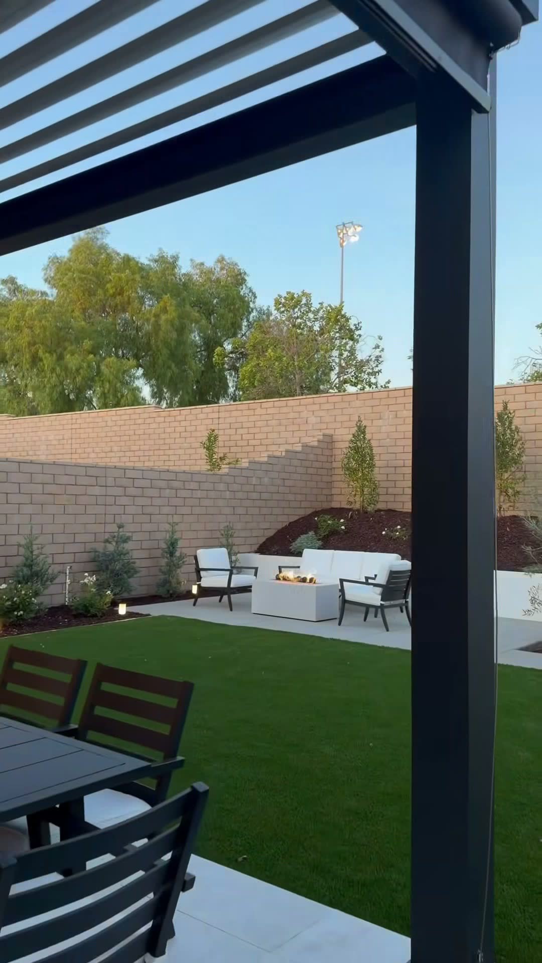 Lit rectangular gas fire pit with white lounge seating on turf, viewed from under a louvred pergola