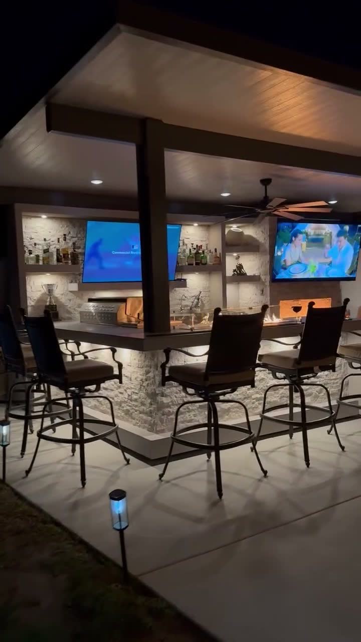 Night view of an L-shaped stone bar with two mounted TVs, a fire trough and swivel bar stools