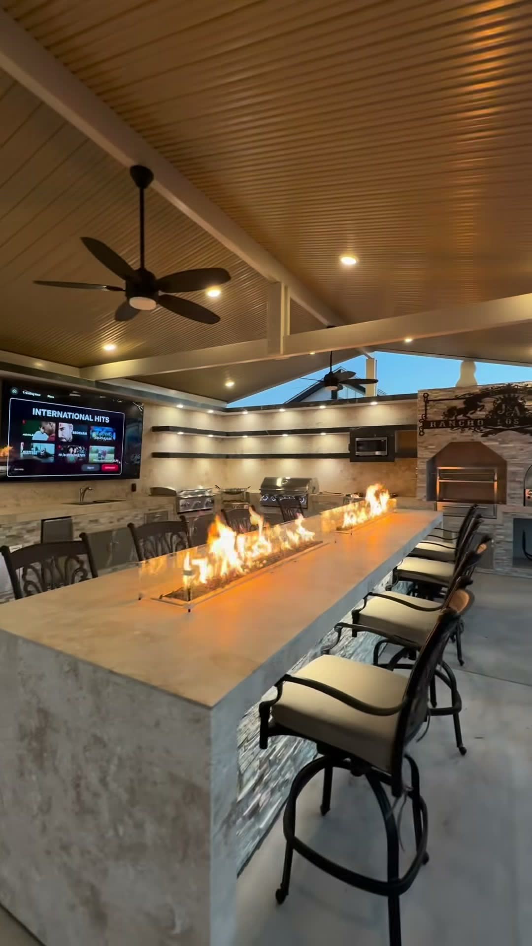 Long linear fire trough burning down a stone bar top under a gable patio cover at dusk