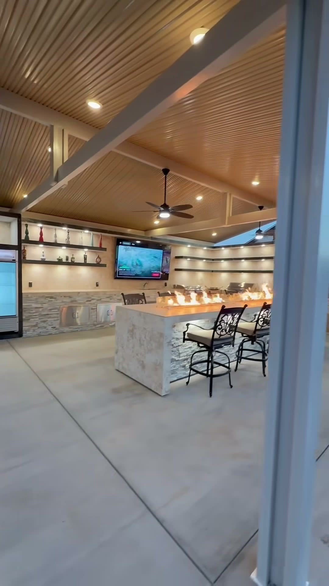 Wide view of a fire-trough bar, mounted TV and beverage cooler under a beamed gable patio cover