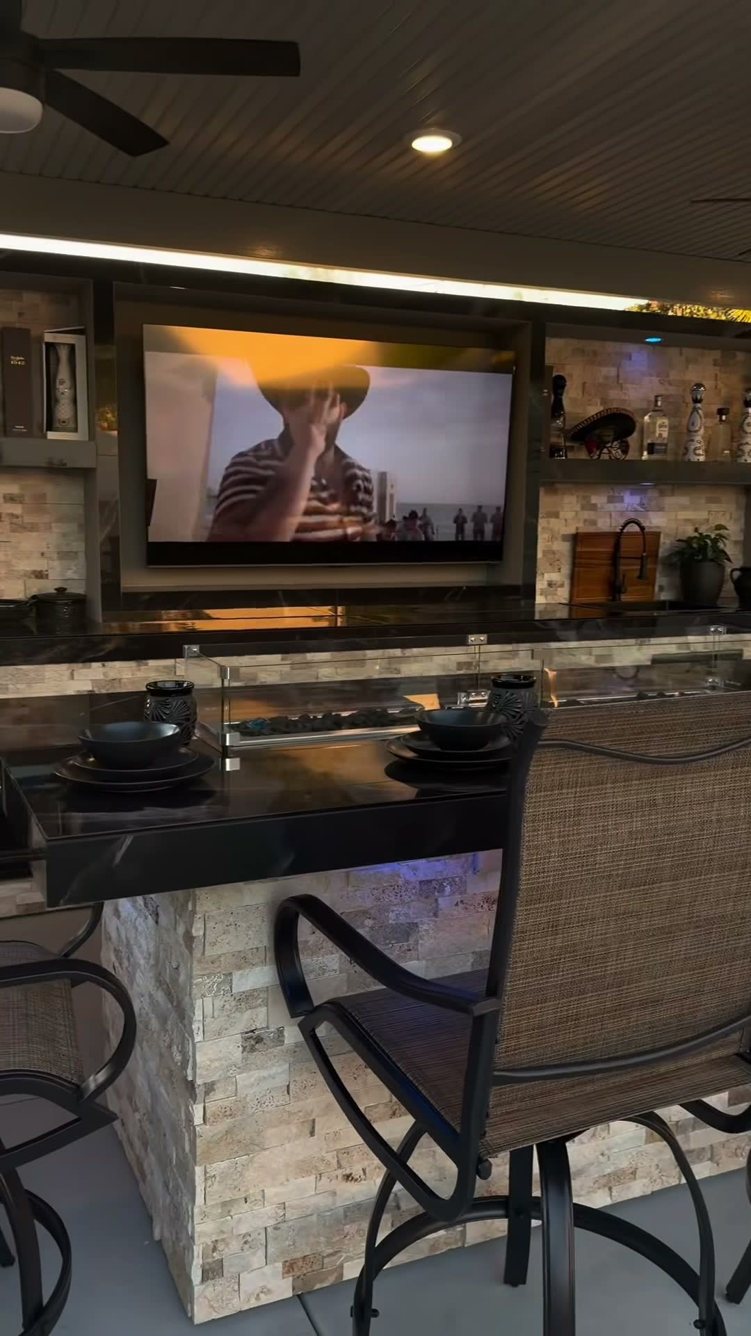 Travertine bar with a glass fire-trough surround below a recessed TV wall and lit liquor shelves