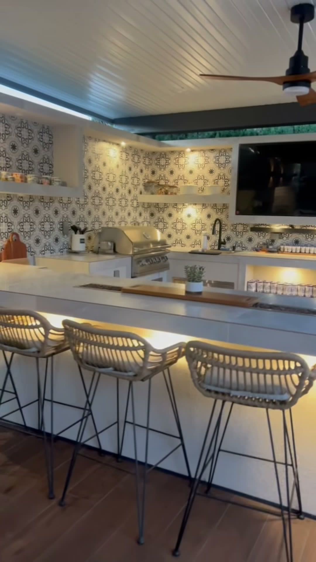 White outdoor kitchen with patterned tile backsplash, stainless grill, mounted TV and LED-lit bar counter