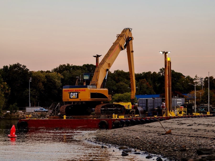 An excavator working from a barge alongside a marina at dusk