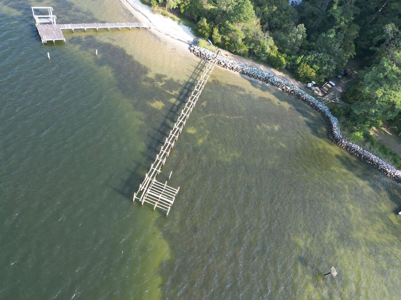 Aerial view along a long timber pier running out from a beach, with a rock revetment armouring the shoreline behind it