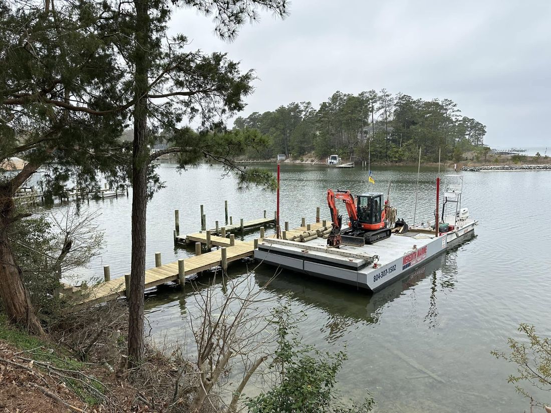The company work barge carrying a red mini-excavator and stacked timbers, moored at the end of a freshly built timber pier