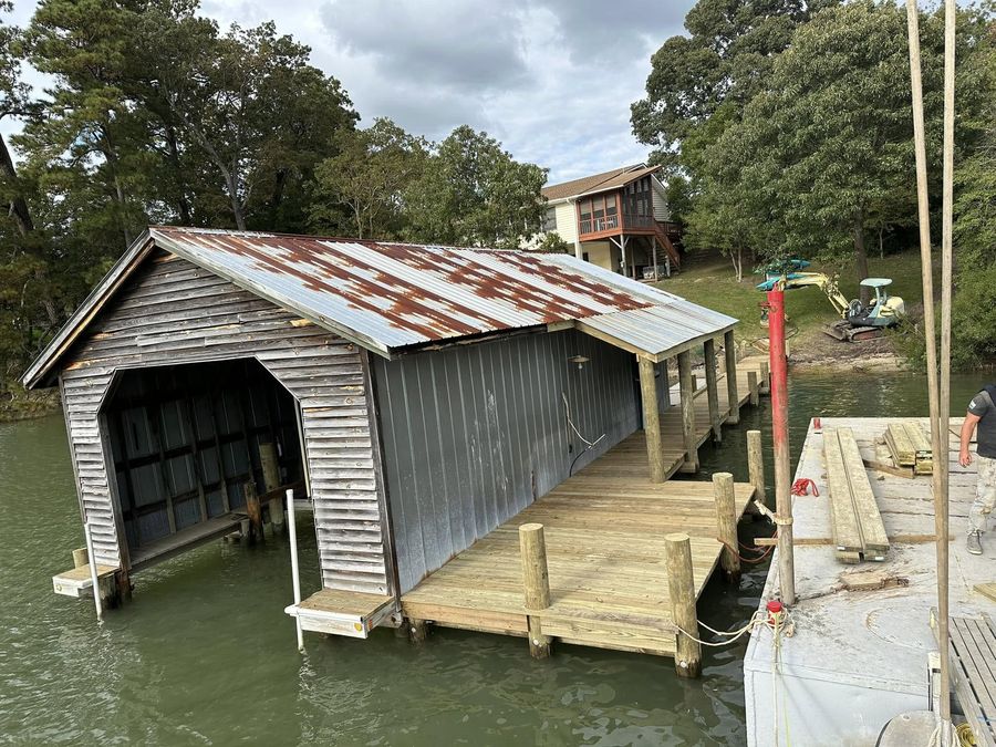 An existing tin-roofed boathouse with new timber decking and pilings built around it, excavator on the bank