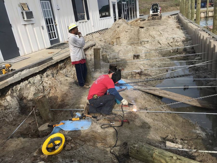 Two crew behind an excavated bulkhead line at Urbanna Marina, one welding a tieback rod running back from the sheet piles