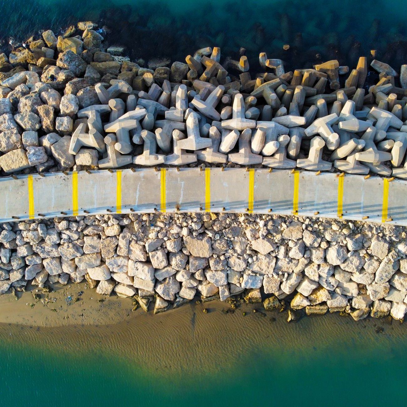 Aerial view of a rock breakwater and its concrete crest walkway meeting turquoise water