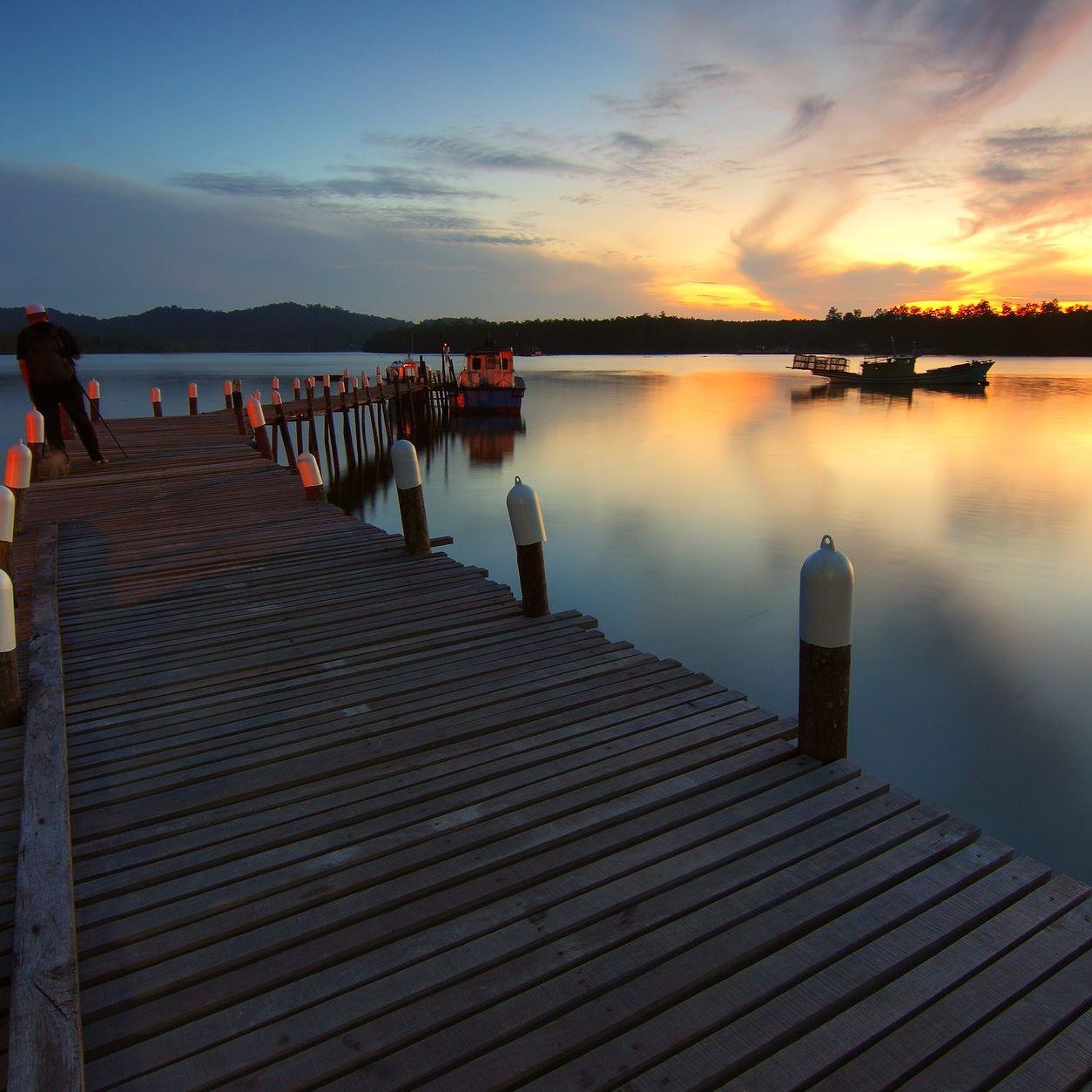 Timber dock running out across still water at sunset, pilings casting long reflections