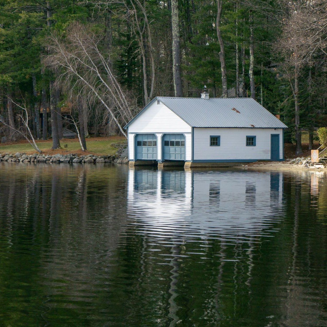 White lakeside boathouse with a pitched roof standing at the edge of the trees
